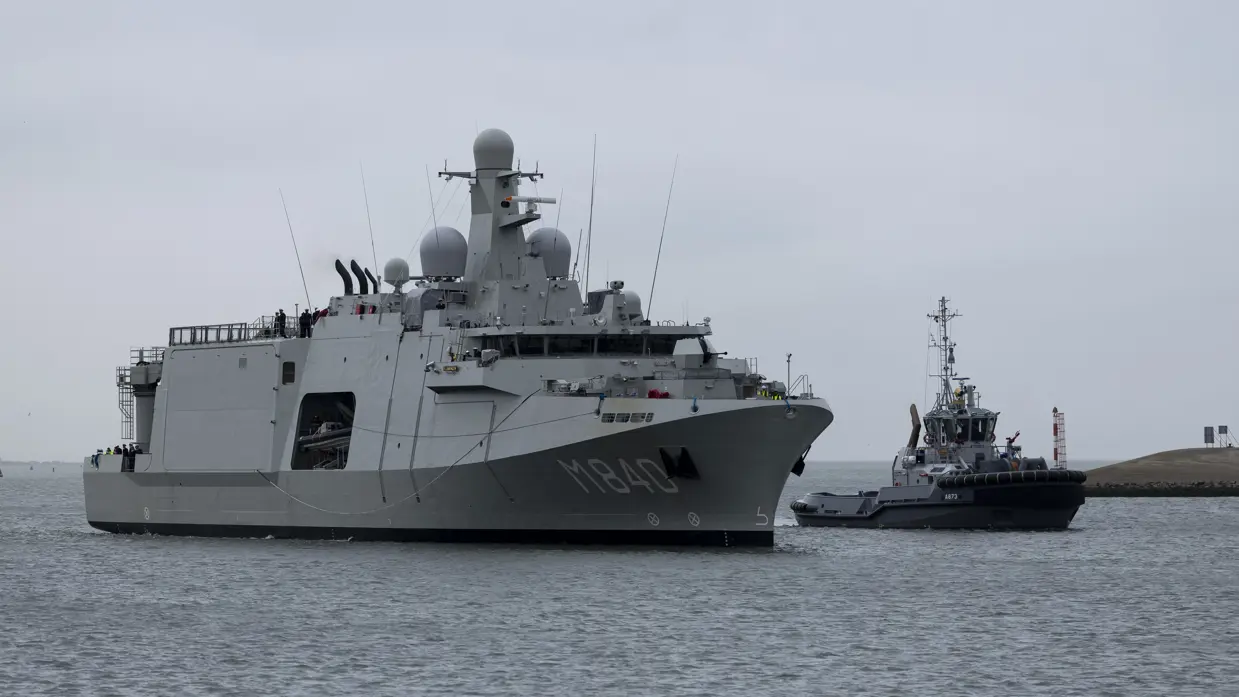 De Vlissingen met daarnaast een sleepboot, gezien vanaf de kade. Daarnaast het grijze marineschip van dichterbij, de sleepboot er nog altijd naast varend.