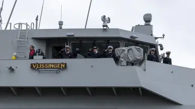: Het varende, grijze marineschip vanaf de zijkant. Daarnaast een afbeelding met daarop een deel van de bemanning bij de reling van het schip, daarachter een wapperende Nederlandse vlag. Vervolgens saluerende militairen vanaf de brug van de Vlissingen en een groepje marineofficieren dat het schip vanaf de kade aanschouwt en vastlegt met de mobiele telefoon.