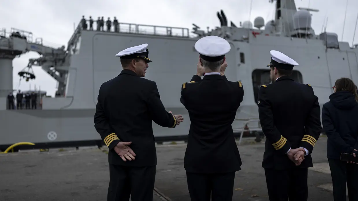 : Het varende, grijze marineschip vanaf de zijkant. Daarnaast een afbeelding met daarop een deel van de bemanning bij de reling van het schip, daarachter een wapperende Nederlandse vlag. Vervolgens saluerende militairen vanaf de brug van de Vlissingen en een groepje marineofficieren dat het schip vanaf de kade aanschouwt en vastlegt met de mobiele telefoon.
