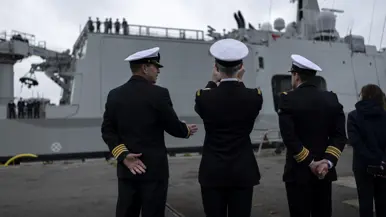 : Het varende, grijze marineschip vanaf de zijkant. Daarnaast een afbeelding met daarop een deel van de bemanning bij de reling van het schip, daarachter een wapperende Nederlandse vlag. Vervolgens saluerende militairen vanaf de brug van de Vlissingen en een groepje marineofficieren dat het schip vanaf de kade aanschouwt en vastlegt met de mobiele telefoon.