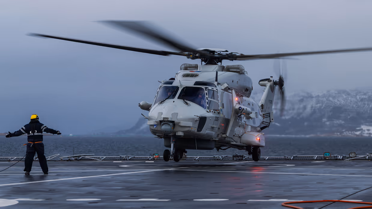 NH90-helikopter landt op het vliegdek van Zr.Ms. Johan de Witt in een Noors fjord bij schemering tijdens oefening Cold Response, terwijl dekpersoneel met lichtsignalen de landing begeleidt.
