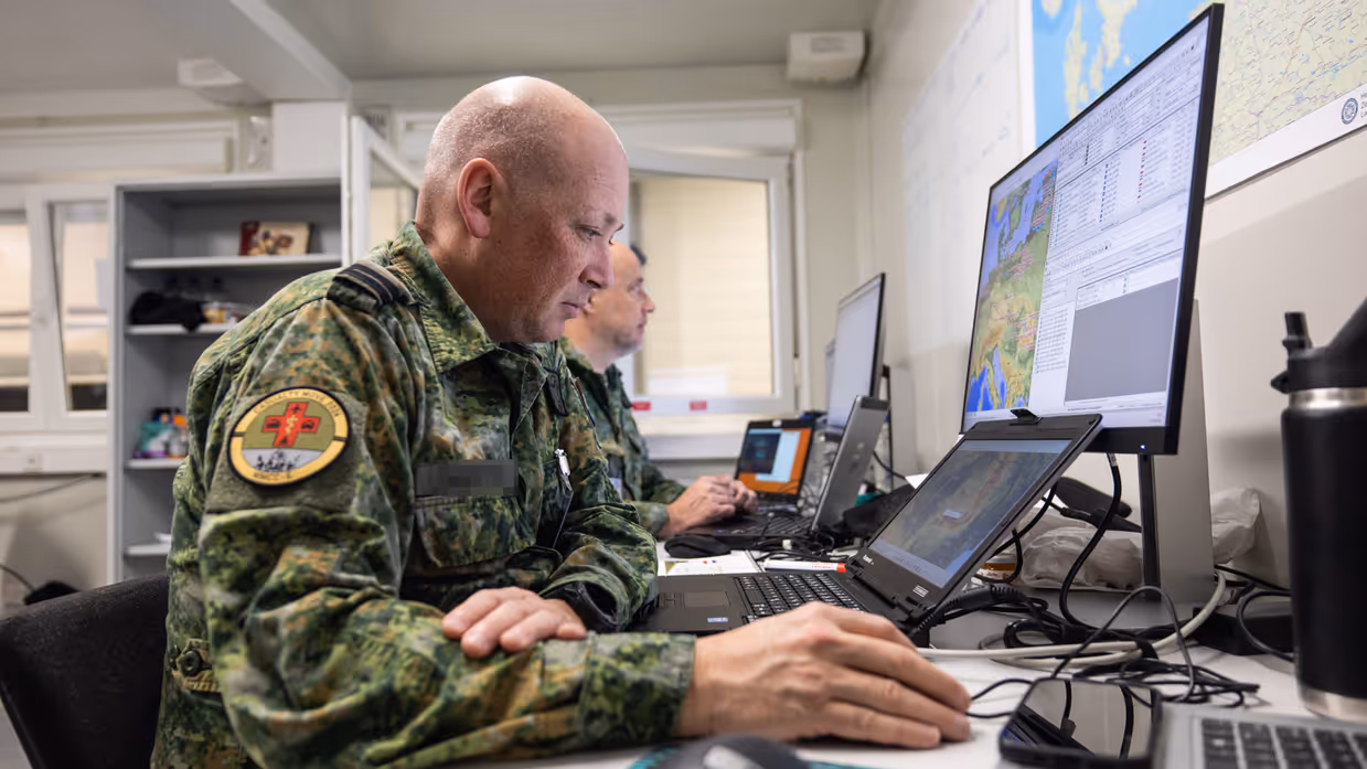 Twee militairen voor meerdere computerschermen aan een bureau. 