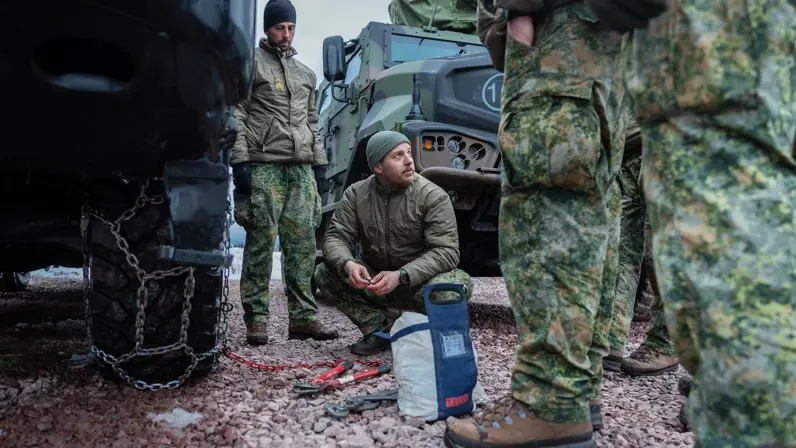Sergeant Timo geeft instructie over het repareren van een sneeuwketting. In zijn hand een grote tang om schakels dicht te knijpen en om hem heen toekijkende manschappen.