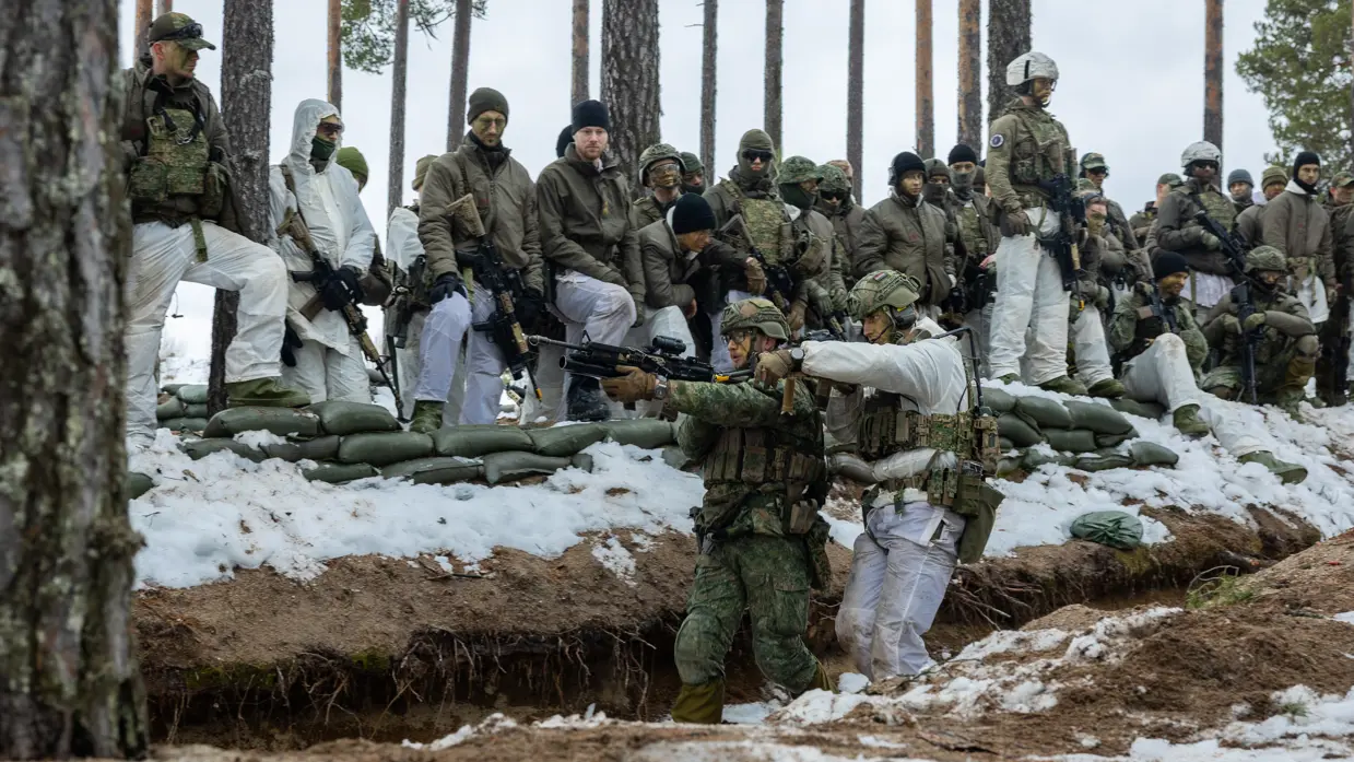 In het bos in winterse omstandigheden geven twee militairen aan collega’ s een demonstratie over verplaatsingen in een loopgraaf.