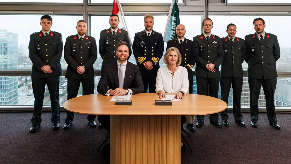 2 mensen zitten aan tafel, achter ze staan reservisten.