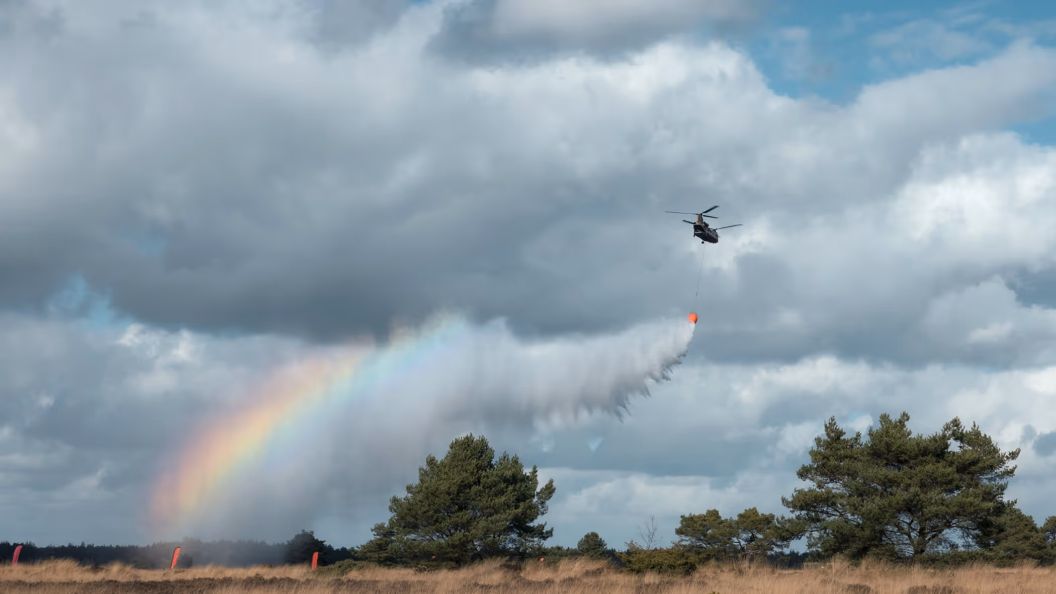 Een helikopter dropt water boven de hei. In de waternevel ontstaat een regenboog.
