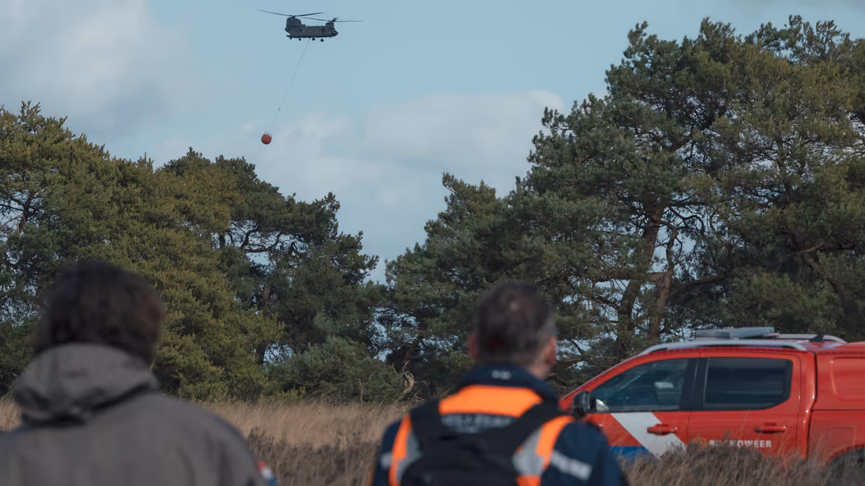 : Een helikopter met Bambi Bucket vliegt tussen de bomen. In de voorgrond lopen een militair en een brandweerman. Rechts staat een rode terreinwagen van de brandweer.