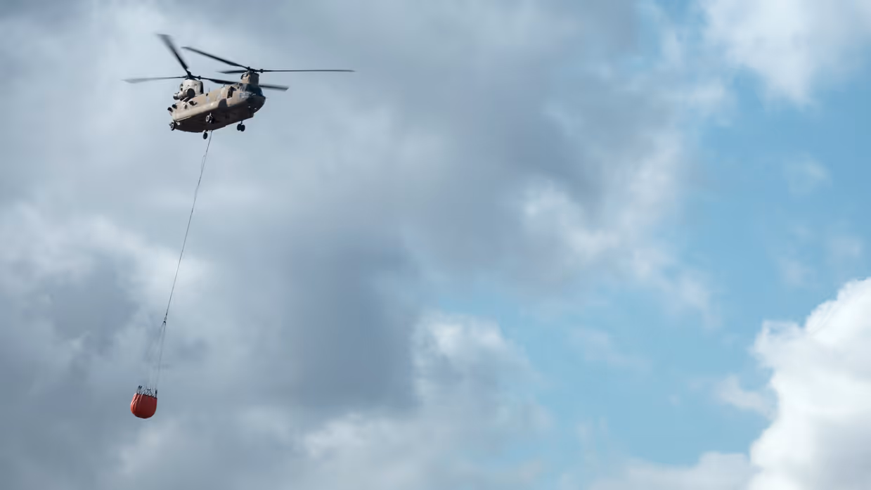 Helikopter met Bambi Bucket komt van links aanvliegen tegen een bewolkte lucht waar de zon op schijnt.