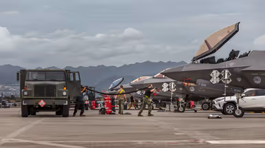 Luchtmachtpersoneel sjouwt brandstofslangen uit een Amerikaanse tankwagen naar een F-35. In de achtergrond zijn bergen te zien.