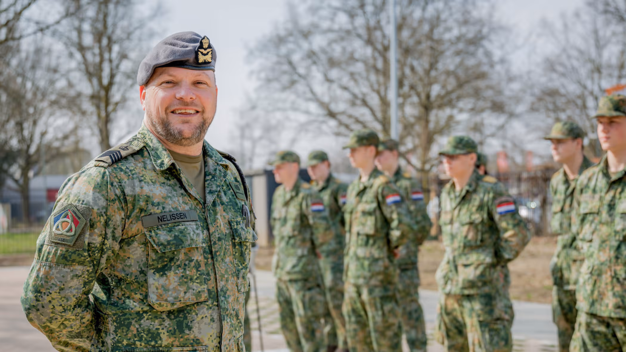 Sergeant-majoor Tim Nelissen poseert buiten voor twee rijen opgestelde militairen met militaire petjes op.