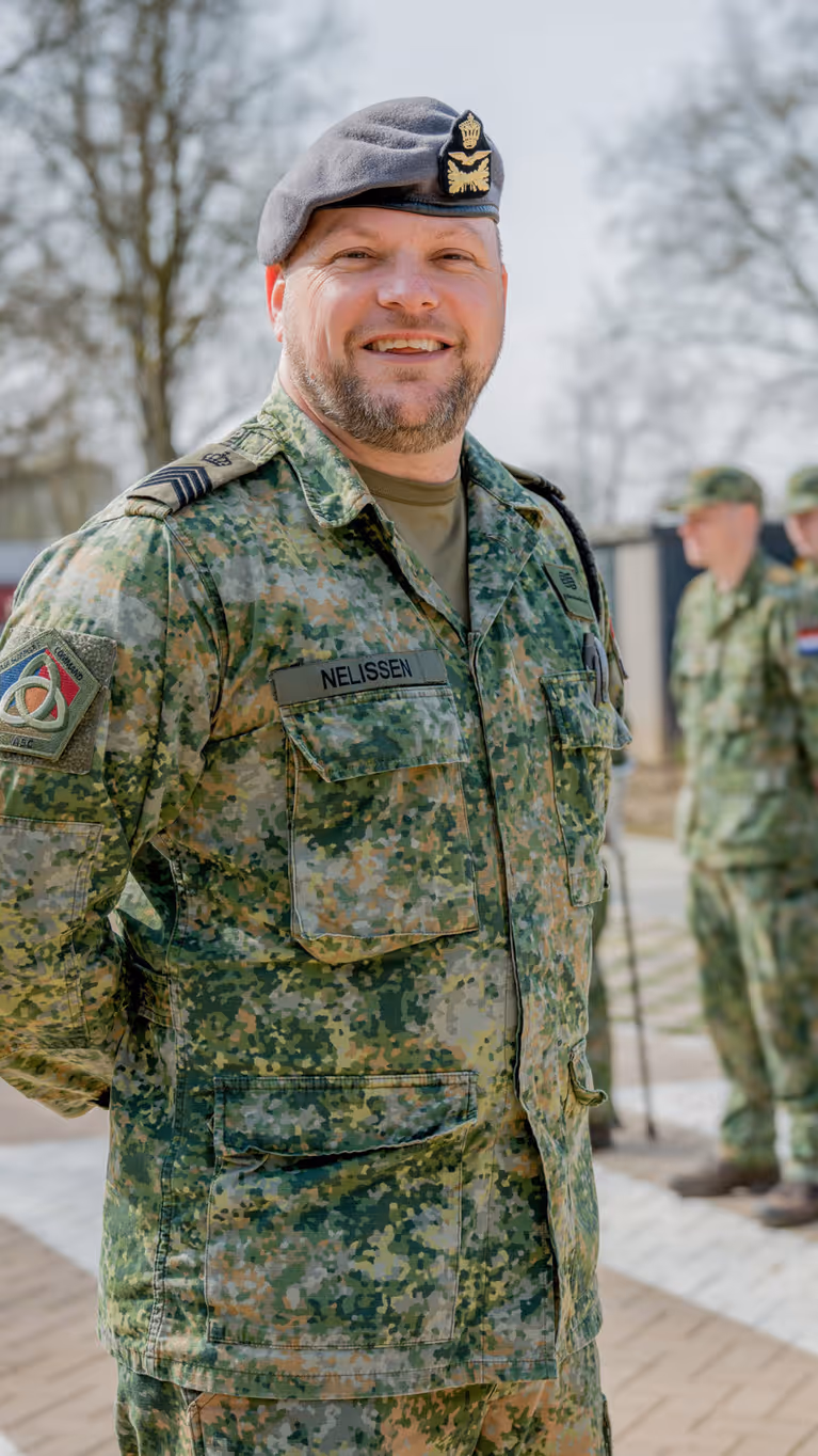 Sergeant-majoor Tim Nelissen poseert buiten voor twee rijen opgestelde militairen met militaire petjes op.