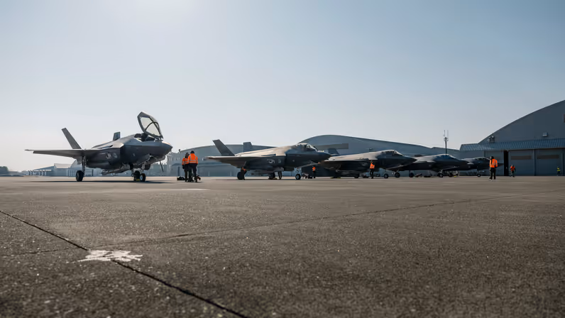 Alt-tekst: 5 F-35’s naast elkaar op een platform met in de achtergrond een aantal ronde hangers. Er staat personeel met oranje jassen tussen de F-35’s. 