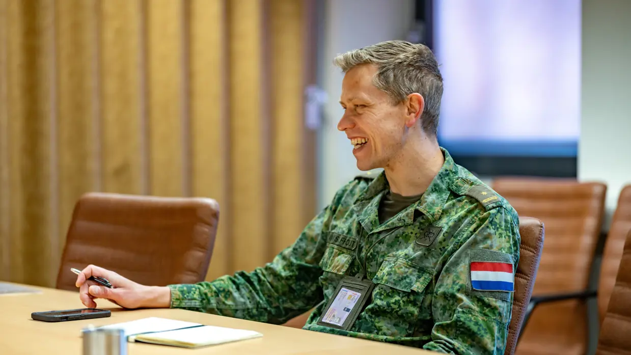 Een militair met pen in hand lacht aan een vergadertafel.
