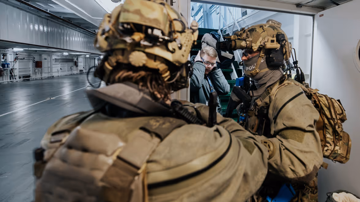  Twee operators van NLMARSOF houden een scheepsdek in de gaten. Op de achtergrond zit een man op de trap die zijn handen over zijn oren heeft. 