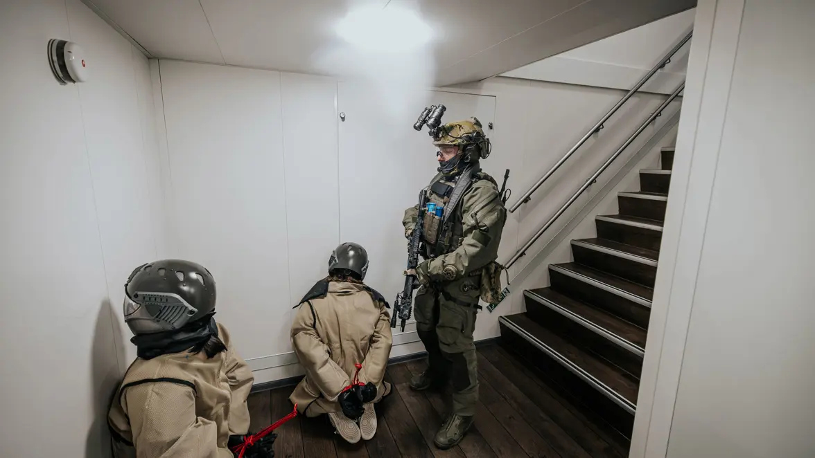 Een operator van NLMARSOF bewaakt twee gevangenen die geknield op de grond zitten, neus richting de muur.