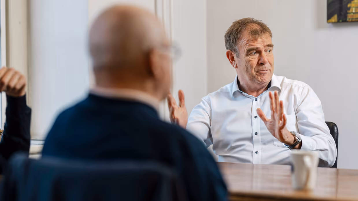 Twee mannen zitten aan een tafel in een lichte kamer. De man rechts, in wit overhemd, praat met handgebaren. Achter hem een raam. 