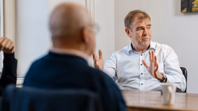 Twee mannen zitten aan een tafel in een lichte kamer. De man rechts, in wit overhemd, praat met handgebaren. Achter hem een raam. 