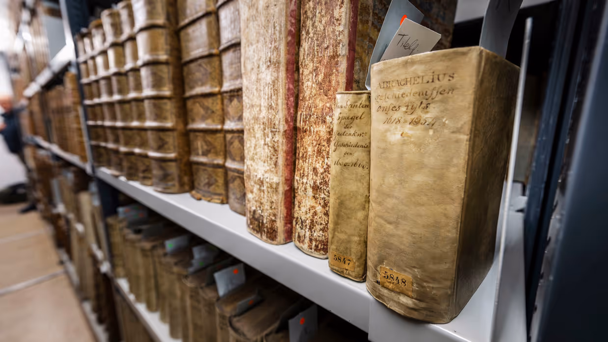Een witte boekenplank met oude, bruine boeken met versleten kaften, sommige met handgeschreven etiketten en nummers.