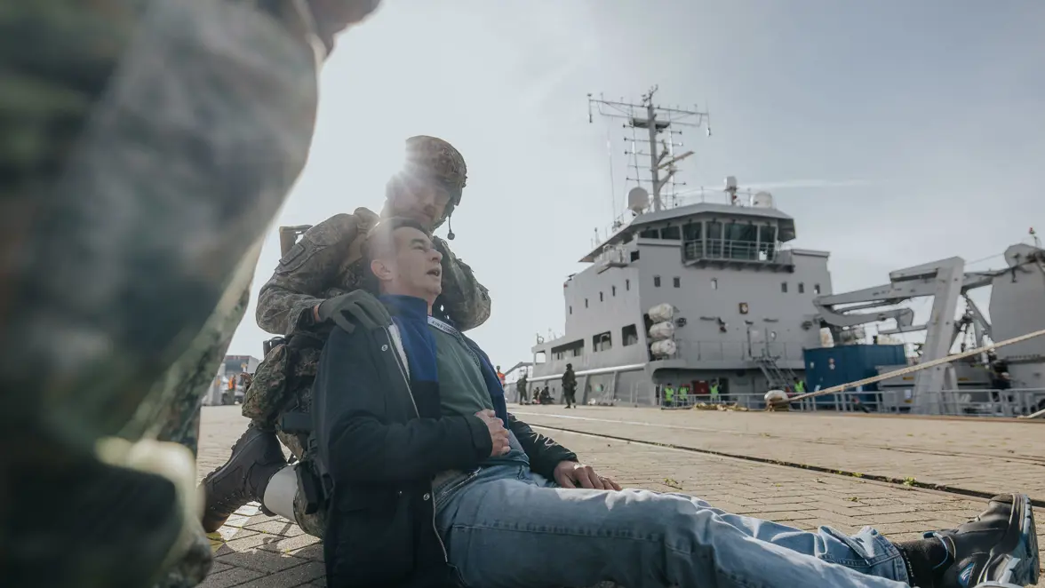 Een militair in groen vlekkenpak ondersteunt een gewonde burger. Op de achtergrond is Zr.Ms. Luymes te zien, aangemeerd aan de kade.  