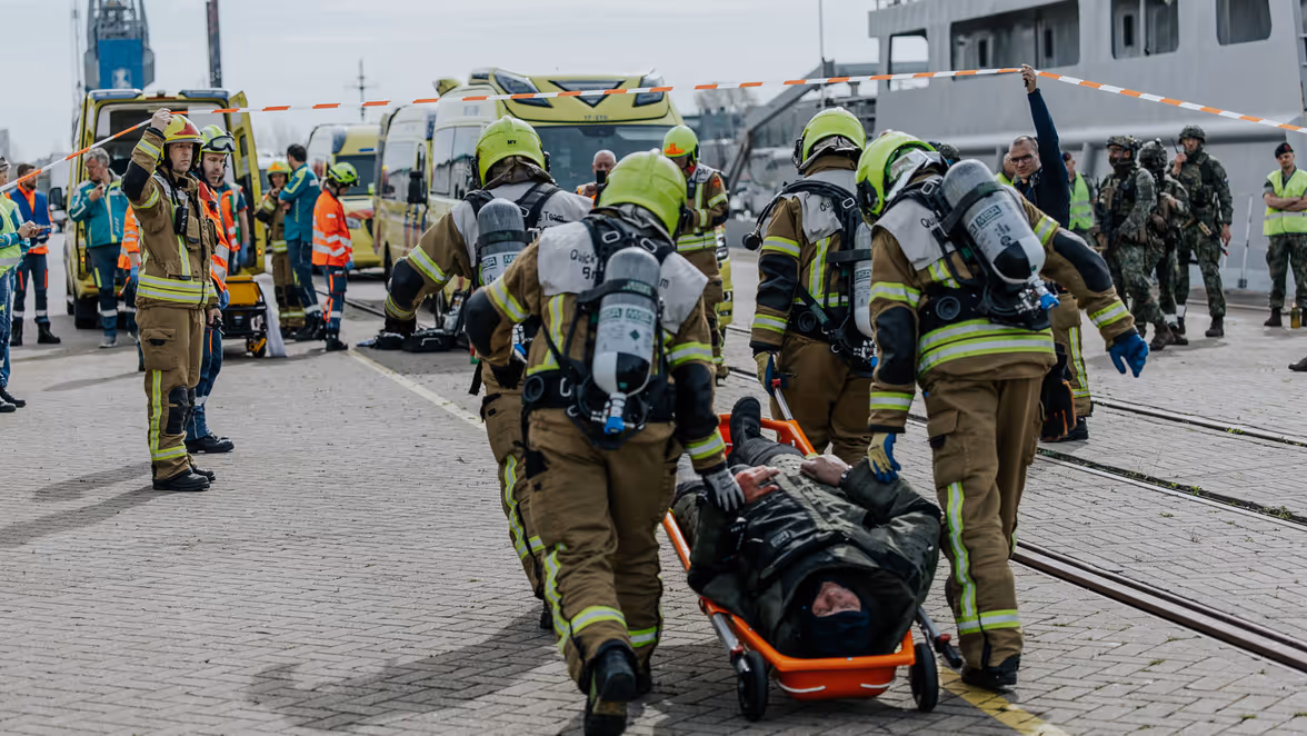 Een man in bompak wordt op een rijdende brancard afgevoerd door brandweerpersoneel. Daarachter is een menigte aan veiligheidspersoneel te zien, waaronder ambulancepersoneel en militairen. 