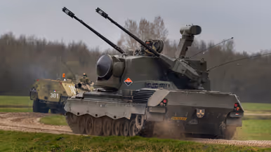 Een tank en gepantserde voertuigen rijden over een onverhard pad door een groen, open veld onder een bewolkte lucht. Rechts: Een zwaar militair voertuig met dubbele kanonnen en radar rijdt over een onverhard pad, gevolgd door andere legervoertuigen in een groen veld.