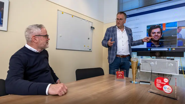 Een man staat bij een televisiescherm en wijst naar een foto van een stagiair. Links van hem zit een man te luisteren naar de uitleg. 