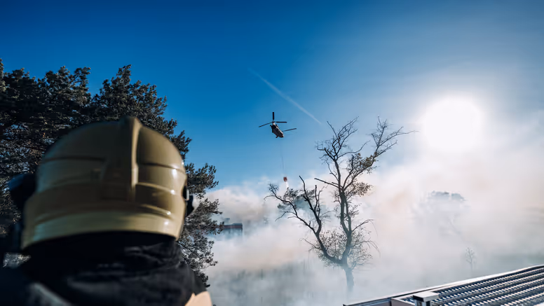 Brandweer blust brand terwijl in de lucht een Chinook-blushelikopter water dropt. 