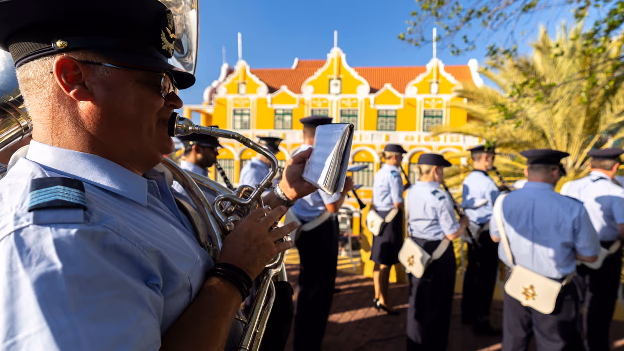 Luchtmachtmuzikanten treden op tegen een achtergrond van gele huizen en palmbomen.
