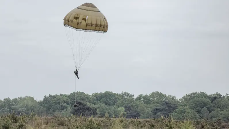 Een parachutist daalt met geopende parachute af boven de Belgische heide terwijl een vrachtlading onder hem hang.