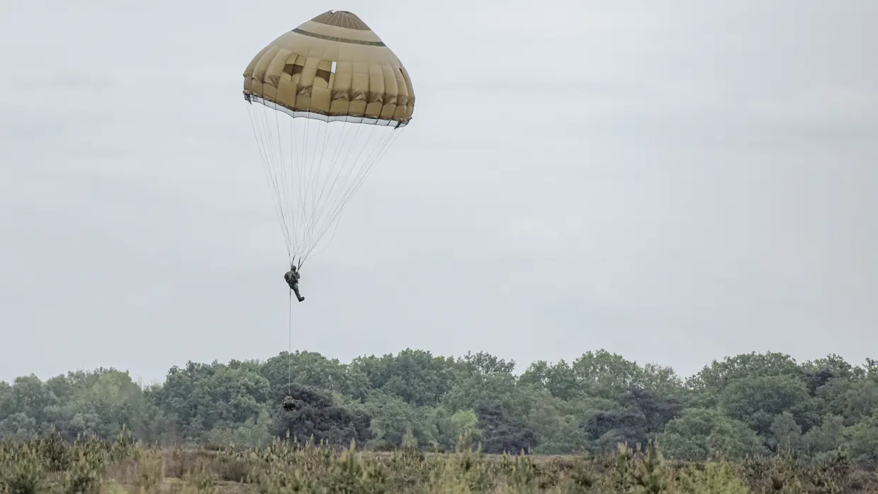Een parachutist daalt met geopende parachute af boven de Belgische heide terwijl een vrachtlading onder hem hang.