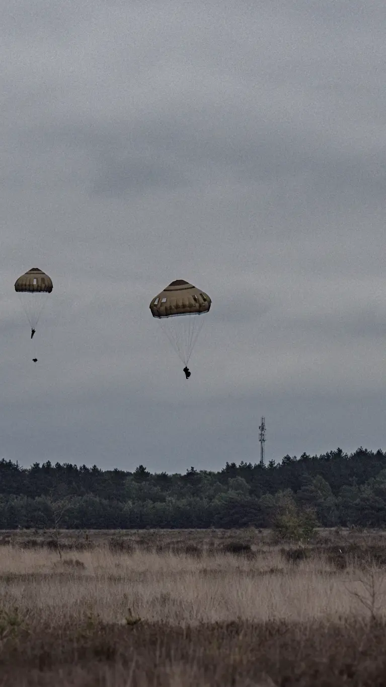 Meerdere parachutisten dalen neer boven de Belgische heide terwijl een transportvliegtuig op de achtergrond wegvliegt tijdens oefening Versatile Guard.