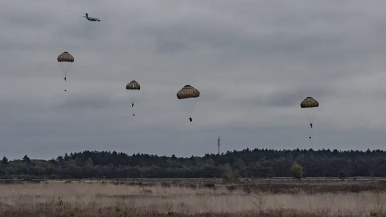 Meerdere parachutisten dalen neer boven de Belgische heide terwijl een transportvliegtuig op de achtergrond wegvliegt tijdens oefening Versatile Guard.