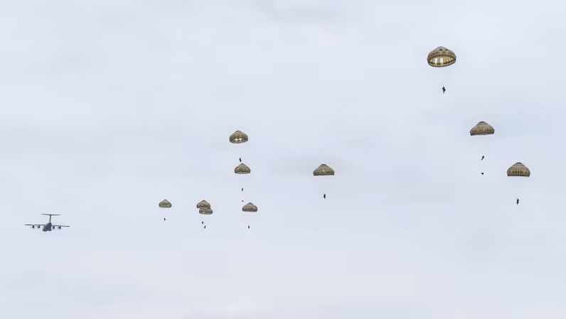 Tien parachutisten in de lucht met op de achtergrond een wegvliegend transportvliegtuig.