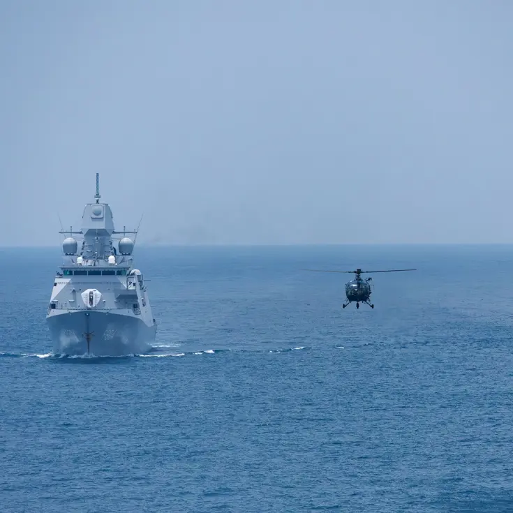 Tussen 2 marineschepen op zee vliegt een helikopter laag boven het water.