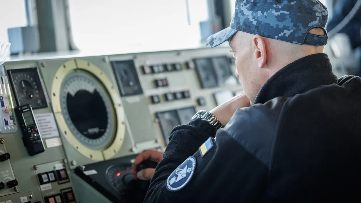 Een militair op de brug, een militair kijkt aan dek door een verrekijker en een militair bedient apparatuur op de brug.