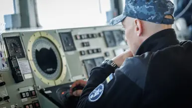 Een militair op de brug, een militair kijkt aan dek door een verrekijker en een militair bedient apparatuur op de brug.