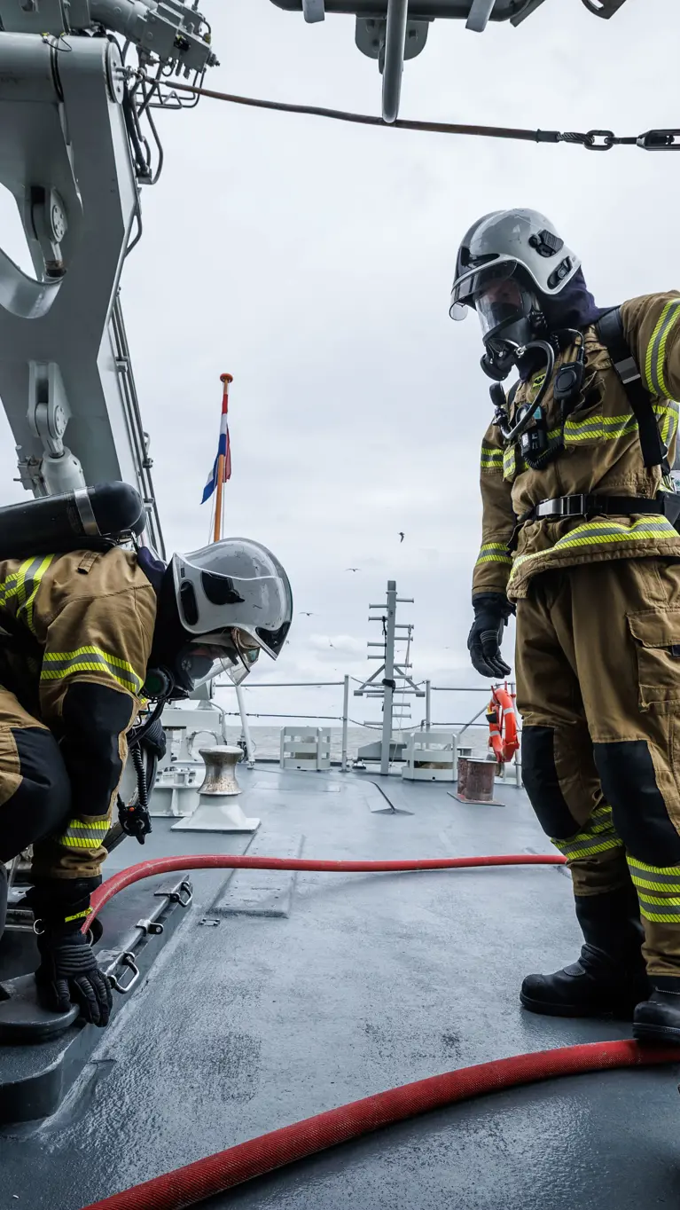 2 brandweermannen in geel pak werken op een marineschip, omringd door metalen constructies en een grijze zee.