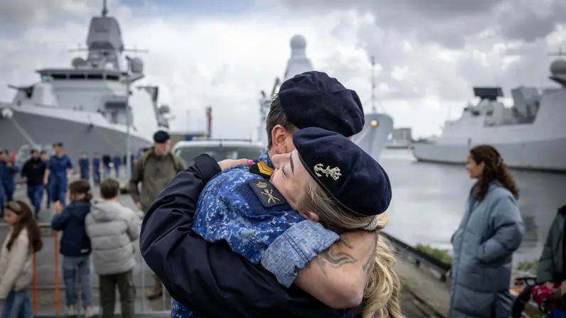 Twee marinecollega’s omhelzen elkaar. Op de achtergrond liggen marineschepen tegen de kade. 