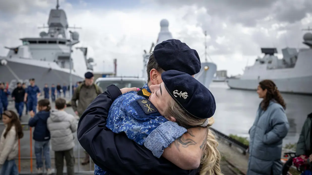 Twee marinecollega’s omhelzen elkaar. Op de achtergrond liggen marineschepen tegen de kade. 