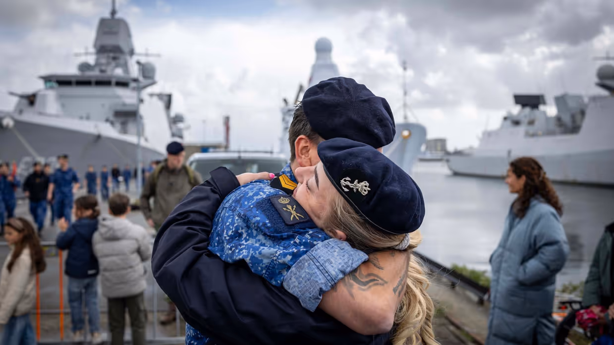 Twee marinecollega’s omhelzen elkaar. Op de achtergrond liggen marineschepen tegen de kade. 