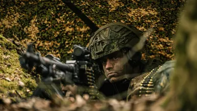 Een militair ligt met een mitrailleur in aanslag onder een poncho. 
