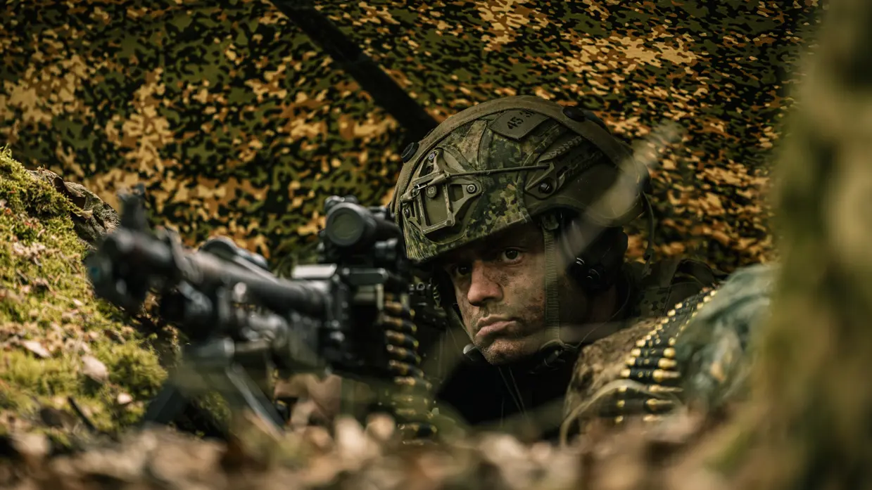 Een militair ligt met een mitrailleur in aanslag onder een poncho. 
