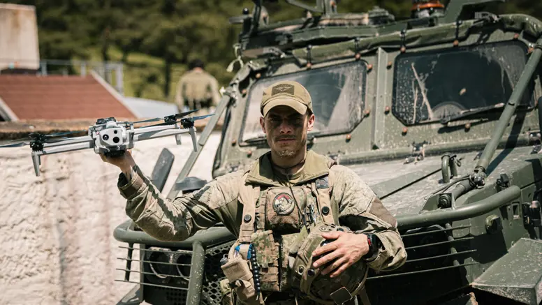 Een militair van de bevoorradingscompagnie van de Luchtmobiele Brigade voor een voertuig met een drone in zijn hand.