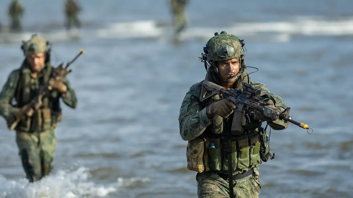 Een marinier loopt door het water, mariniers op het strand, mariniers verdekt opgesteld en een marinier roept bevelen.