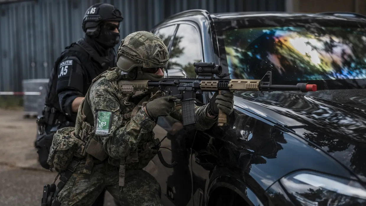Militair richt zijn wapen op een auto. Daarachter staat iemand van de BE.