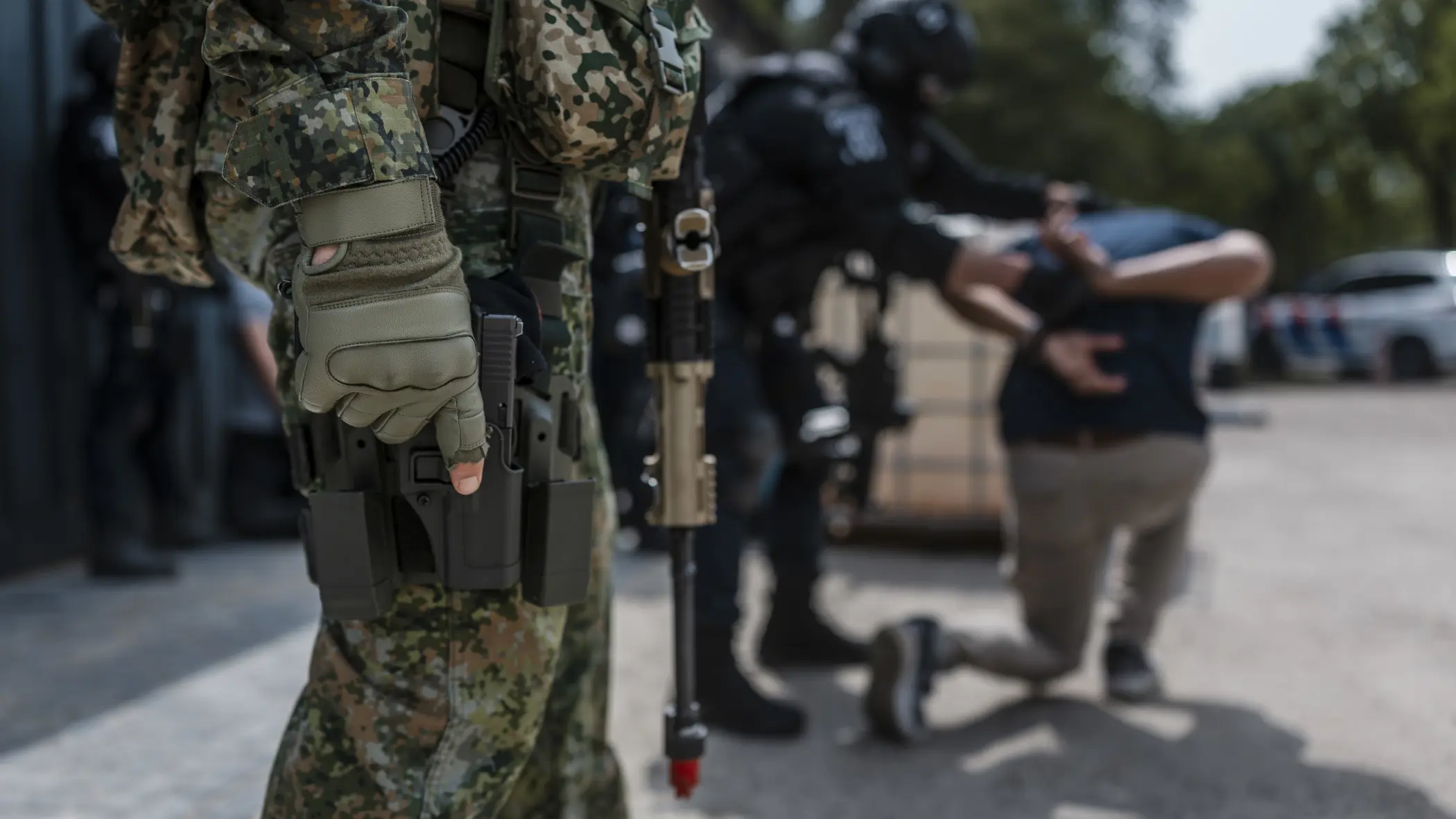 Militair staat op de voorgrond paraat met wapen en op de achtergrond wordt iemand aangehouden door de Politie.