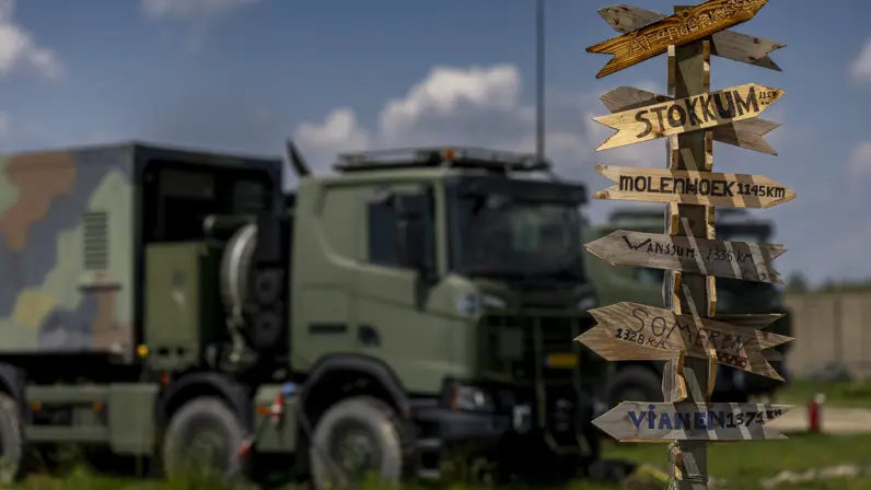 Een paal met daarop allemaal wegwijzers naar plaatsen in Nederland inclusief de afstand. Op de achtergrond staat een vrachtwagen.