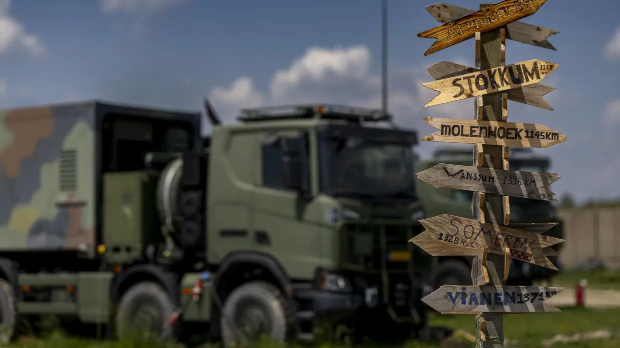 Een paal met daarop allemaal wegwijzers naar plaatsen in Nederland inclusief de afstand. Op de achtergrond staat een vrachtwagen.