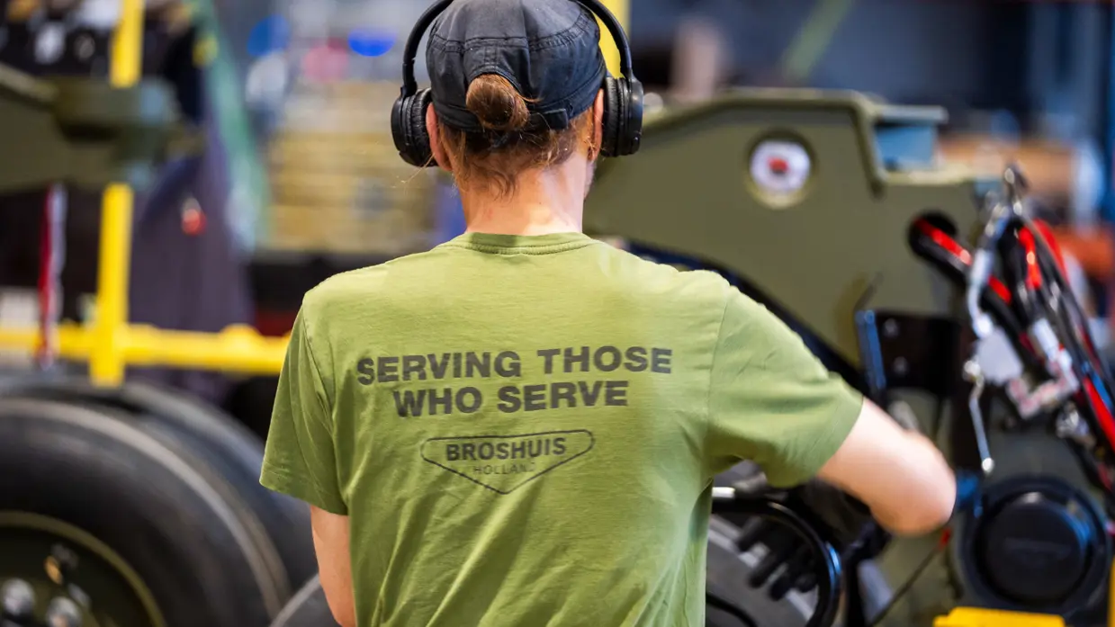 Monteur sleutelt aan wiel in garage en draagt T-shirt met tekst ‘serving those who serve’ op rug.
