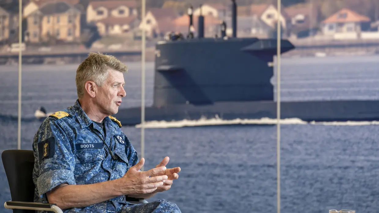 Vice-admiraal Boudewijn Boots praat met beide handen vanuit zijn stoel, voor een poster van een onderzeeboot. 